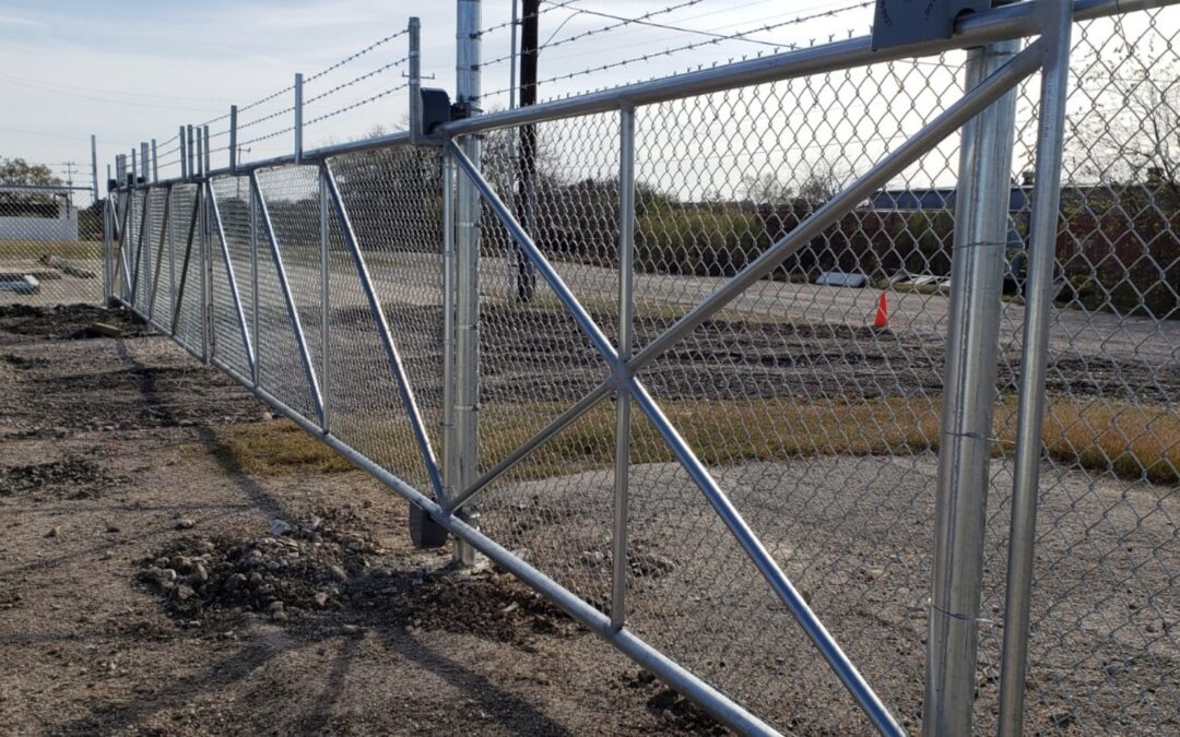 Commercial chain link perimeter fence with a vehicle gate and bollards outside a warehouse.